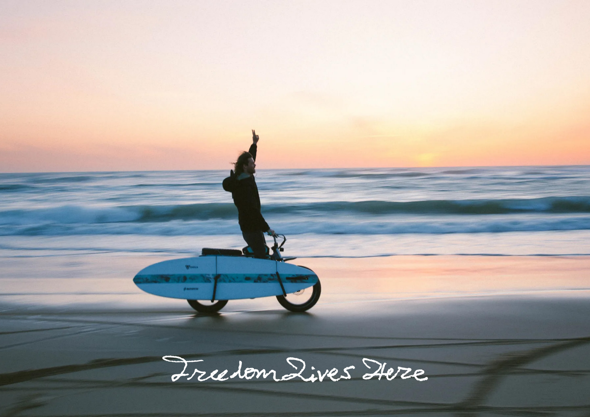 Freedom Lives Here — cyclist carrying a surfboard along the beach at sunset